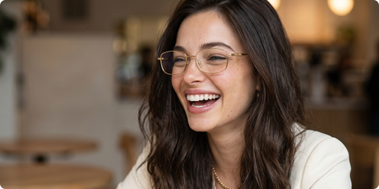 A woman with long, flowing hair and a radiant smile wears rectangular glasses with slender gold frames. She is dressed in a white blazer and a gold necklace, set against the backdrop of a softly lit café.