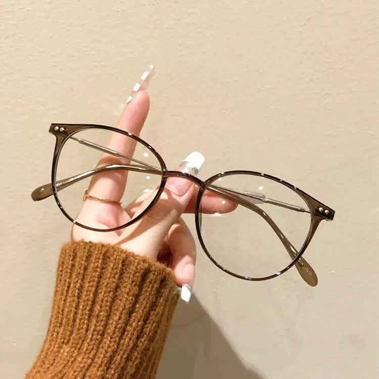 Holding a pair of glasses featuring thin-rimmed, transparent brown resin frames; the lenses are designed with a large, round shape, while the slender temples are accented with decorative gold metal details.