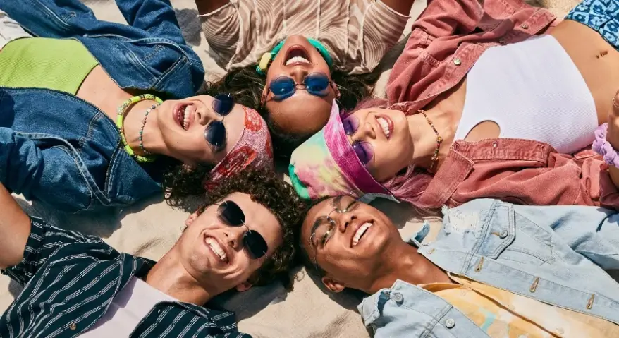 Five young people, wearing sunglasses of various styles, lie in a circle on the beach, beaming with radiant smiles that capture the vibrant atmosphere of a summer outdoors.