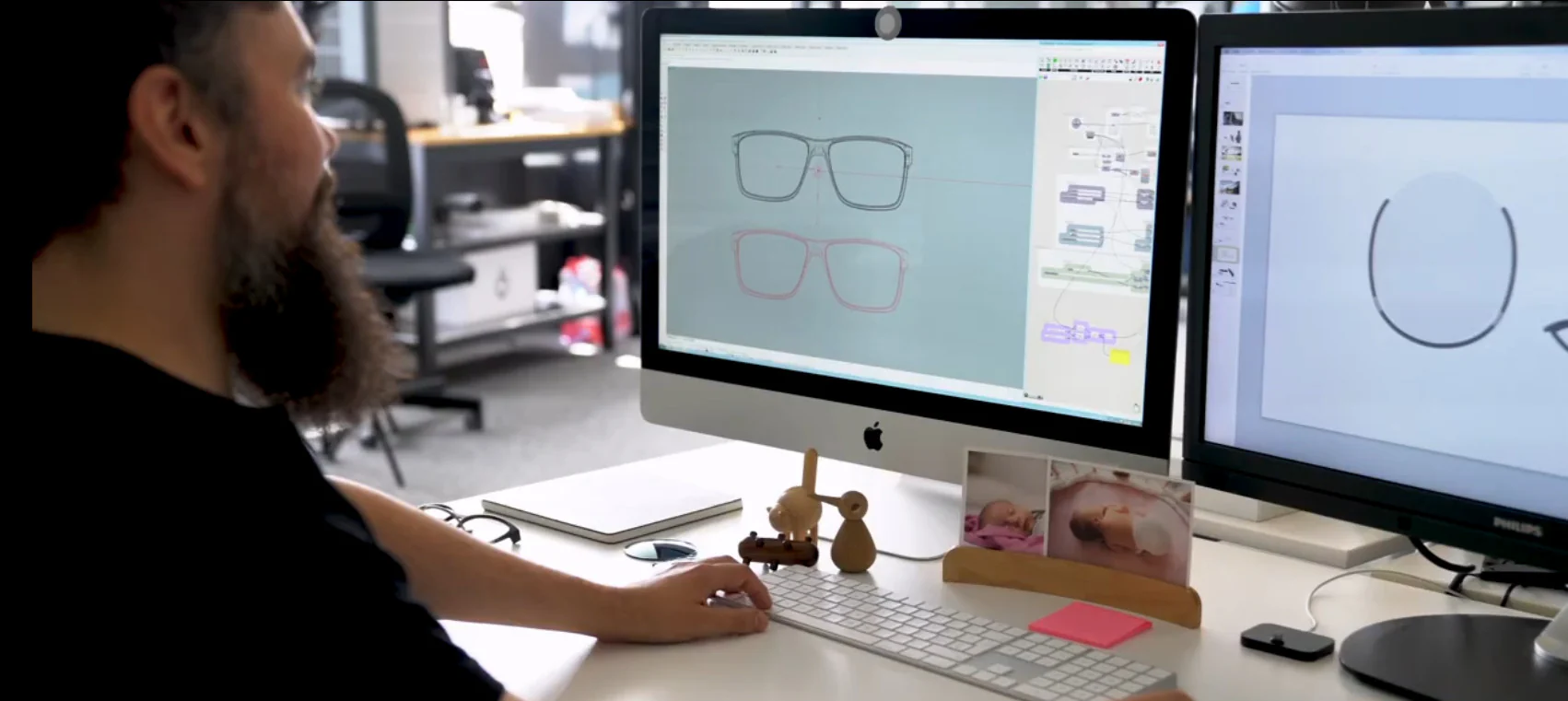 A bearded designer is using a dual-monitor setup to operate CAD software for the 3D modeling of eyeglass frames.