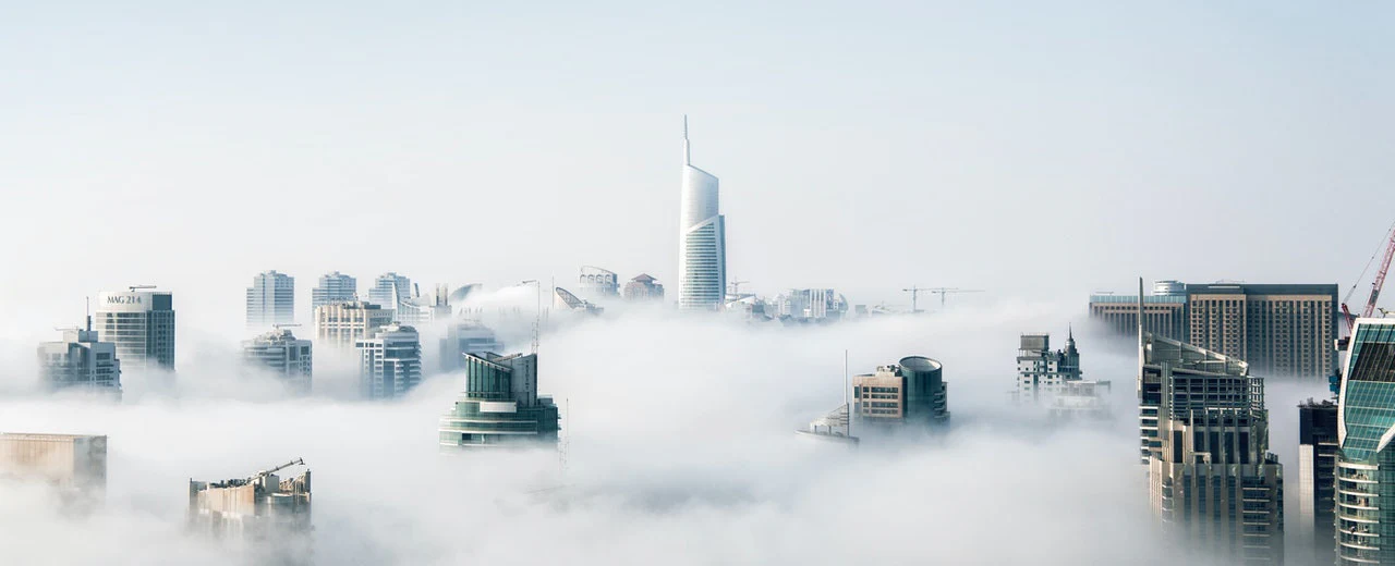 A modern city skyline shrouded in morning mist, with high-rise buildings peeking out from amidst dense white clouds.