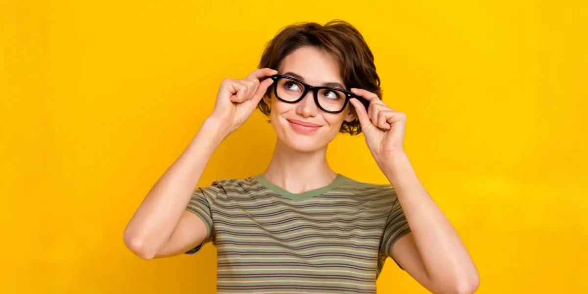 A young woman smiles and pushes up her thick-rimmed black glasses while looking upward against a bright yellow background.