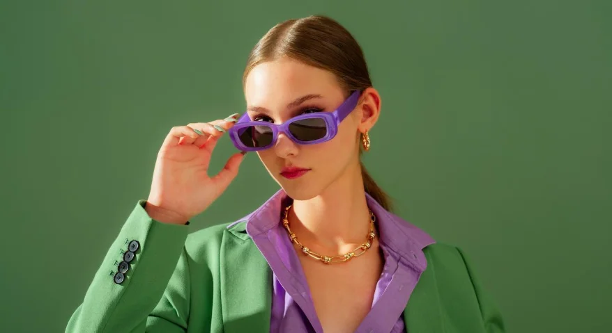 A female model wearing purple-framed sunglasses, a green suit, and a purple shirt, against a green background.