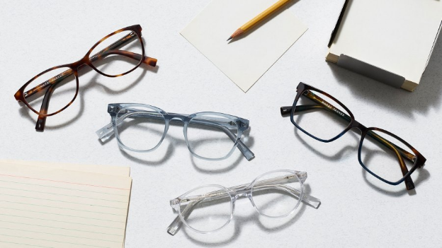 Four pairs of stylish framed glasses scattered across an office desk, with pencils and a notepad in the background.