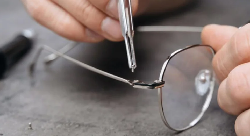 A close-up view of tweezers being used to repair a pair of glasses hinges on a desktop.