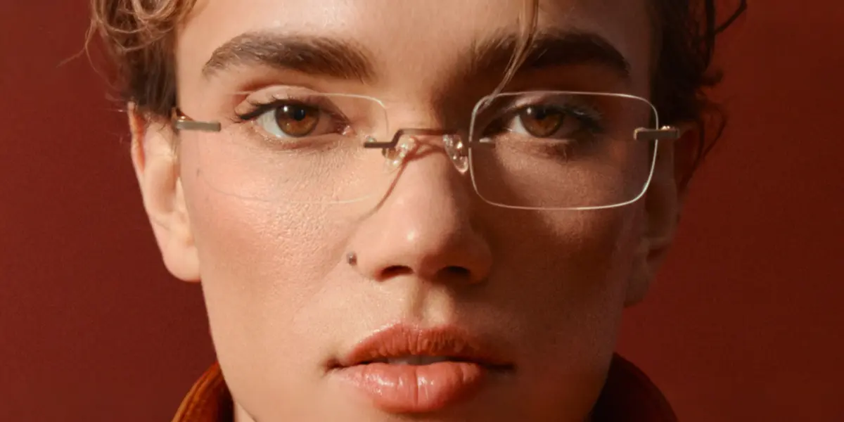 A close-up headshot of a model wearing minimalist, rectangular, rimless glasses, set against a dark red background.