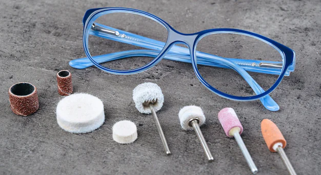 Resting on a gray workbench are a pair of blue eyeglass frames and a variety of rotary grinding heads in various shapes, used for sanding and polishing.