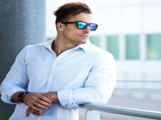 A muscular man wearing a light blue shirt leans outdoors, sporting iridescent blue-mirrored sports sunglasses.