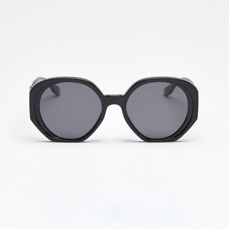 A front view of pure black, oversized sunglasses, featuring a frame design that combines slightly angular hexagonal and circular elements, fitted with dark gray lenses.