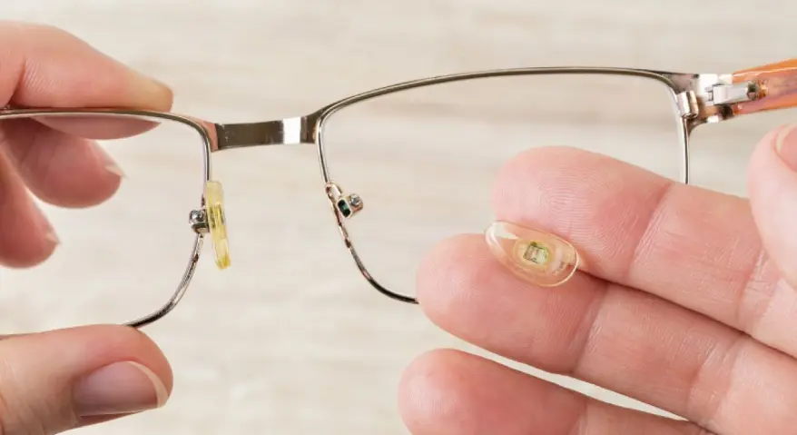 A hand is holding a pair of glasses, showcasing the details of the replaceable or damaged transparent silicone nose pads.