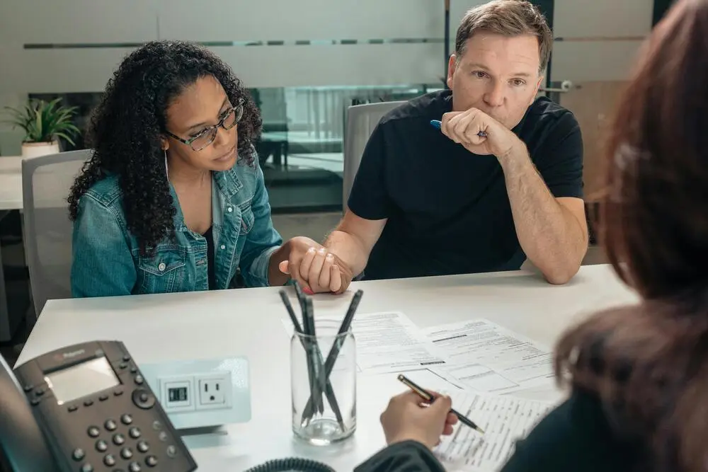 A couple with serious expressions are holding hands at a desk, discussing vision insurance with a professional.