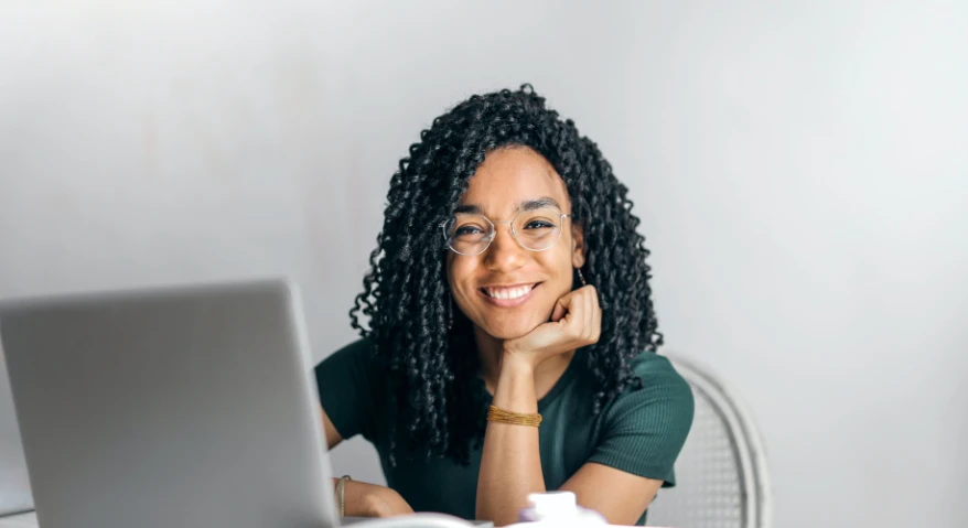 A young woman with dark curly hair, wearing round-rimmed glasses, sits at a computer and smiles confidently at the camera against a simple background.