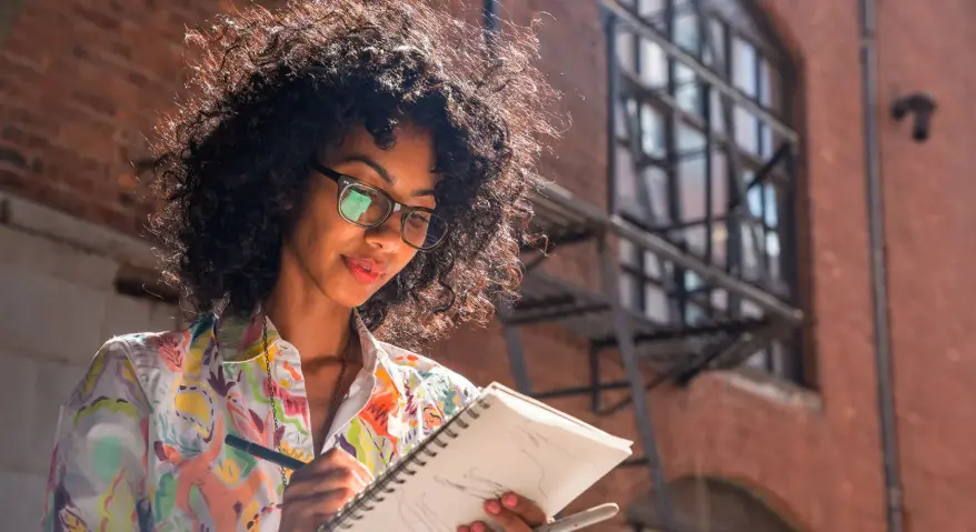 A woman wearing black-rimmed progressive glasses is sketching outdoors in the bright sunshine.