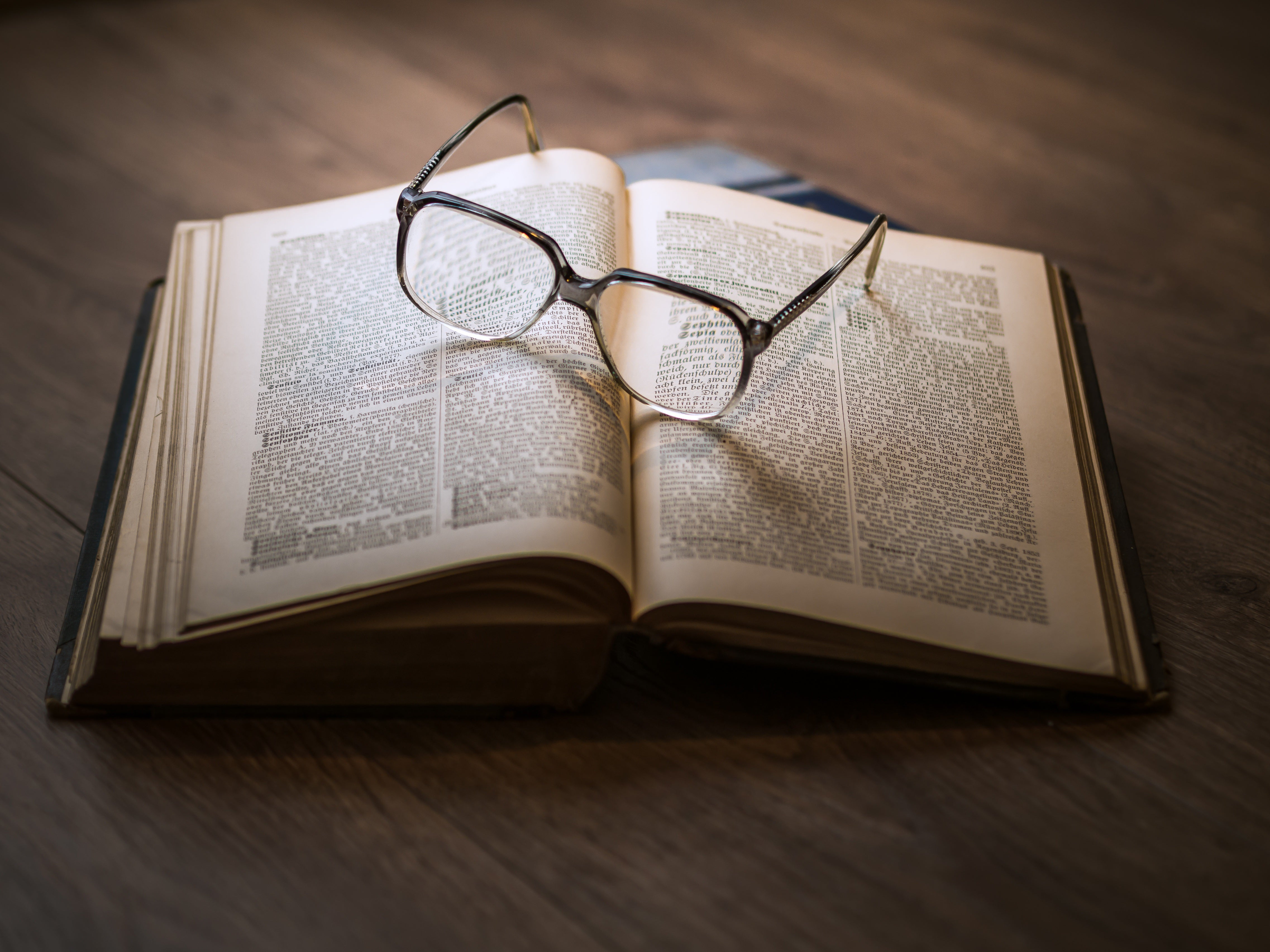 A pair of vintage, thin-rimmed glasses rests upon an open, aged book, evoking a rich academic atmosphere.
