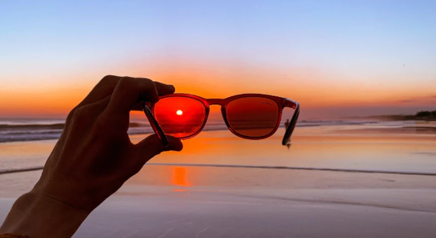 On a beach at sunset, a hand holds up a pair of sunglasses with reddish-brown lenses; viewed through the lenses, the setting sun takes on warmer, more saturated red tones.
