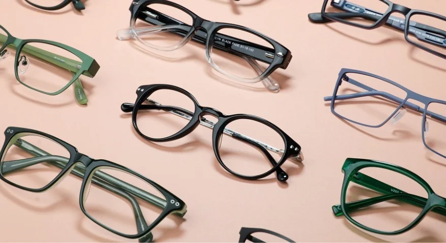 A top-down view of various stylish eyeglass frames—including black round frames, green-and-black square frames, transparent gradient frames, and thin metal-rimmed frames—set against a soft pink background.