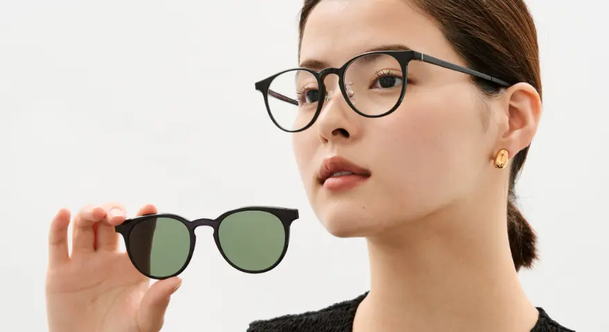A woman wearing black, round-rimmed glasses holds a pair of matching magnetic green sunglass lenses in her hand, demonstrating the magnetic attachment feature.