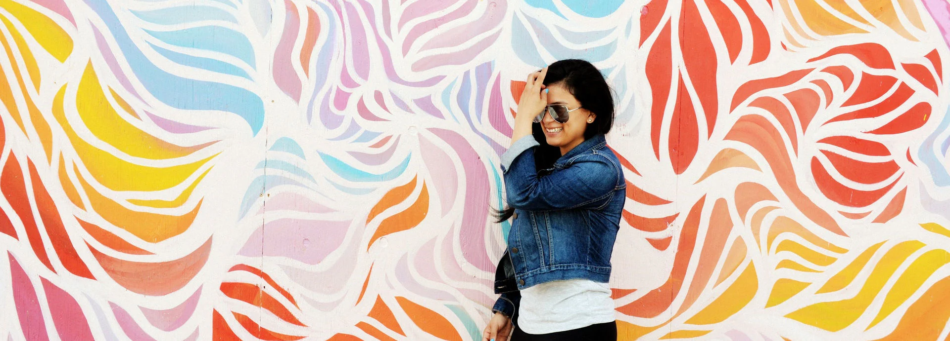 A woman wearing a denim jacket and aviator sunglasses smiles in front of a massive, colorful, streamlined mural.