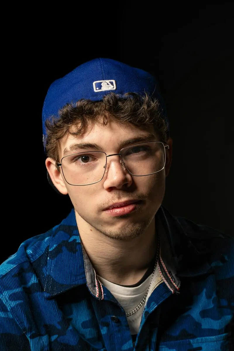 A portrait of a young man wearing a blue baseball cap, black-rimmed glasses, and a blue camouflage shirt.