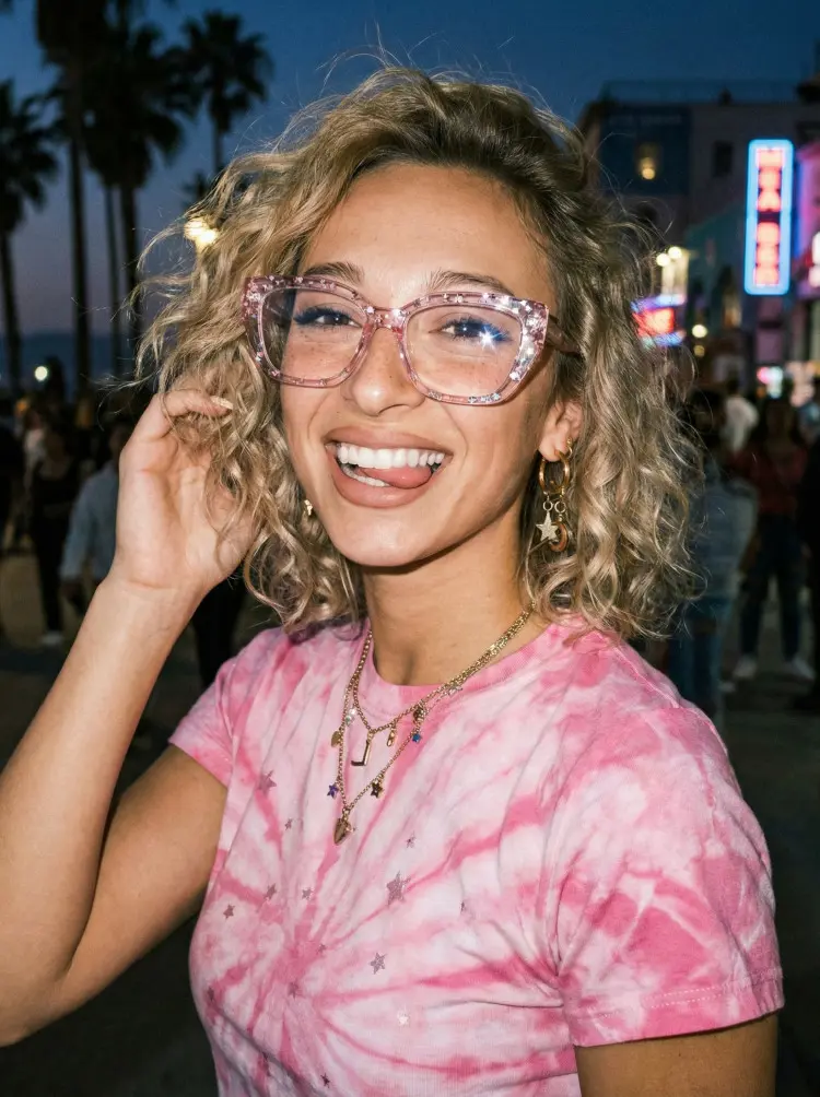 Female model wearing glitter pink cat eye acetate eyeglasses in a pink tie-dye t-shirt smiling on a city street at night with palm trees background. SPU: CA0282601