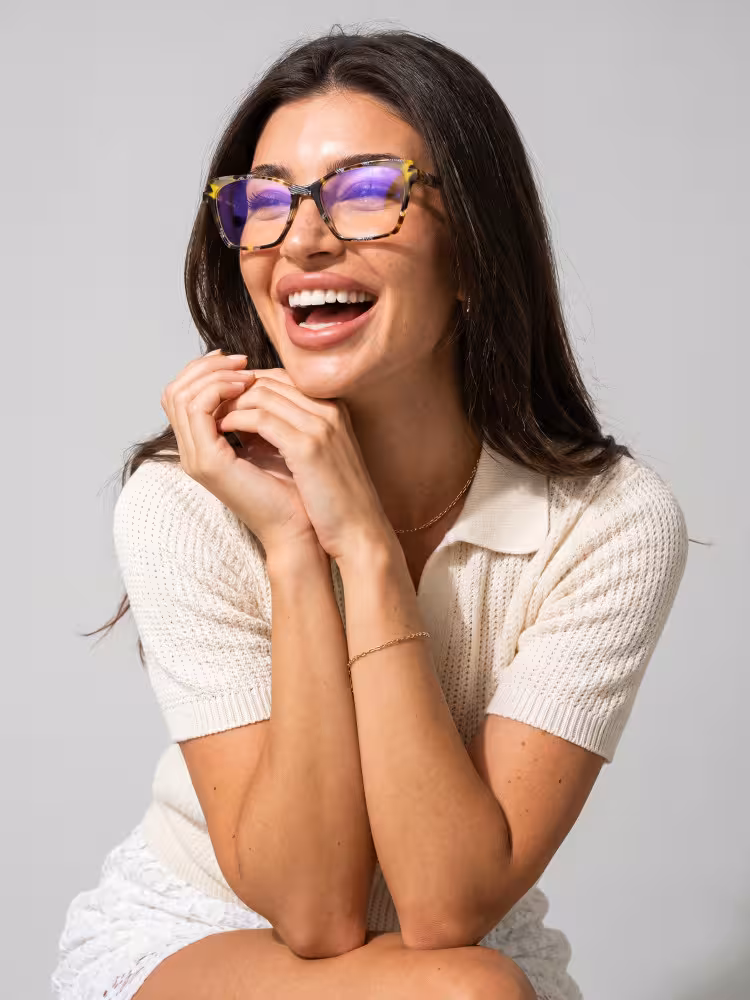 Female model wearing colorful tortoise cat eye acetate eyeglasses in a beige knit polo shirt laughing with hands under chin against a plain grey studio background. SPU: CA0352605