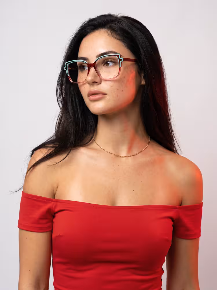Female model wearing colorful two-tone cat eye acetate eyeglasses in a red off-the-shoulder top looking aside against a plain grey studio background. SPU: CA0372605