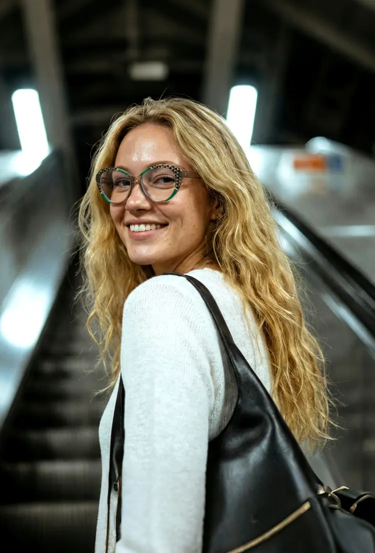Female model wearing everyday black cat eye tr90 eyeglasses in a white knit top with a black leather bag riding an escalator in a subway station. SPU: CR0042405