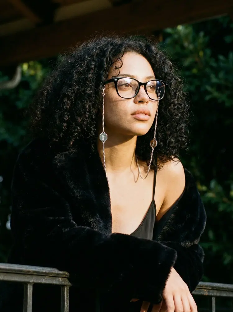 Female model wearing fashion black rectangle tr90 eyeglasses in a black furry coat leaning on a balcony rail with green plants background. SPU: ER0212601