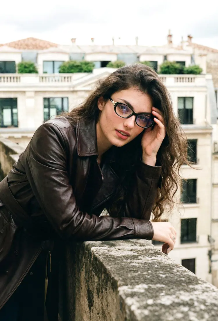Female model wearing office siren black rectangle tr90 eyeglasses leaning on a stone balcony with a classic European city building background. SPU: ER0442509