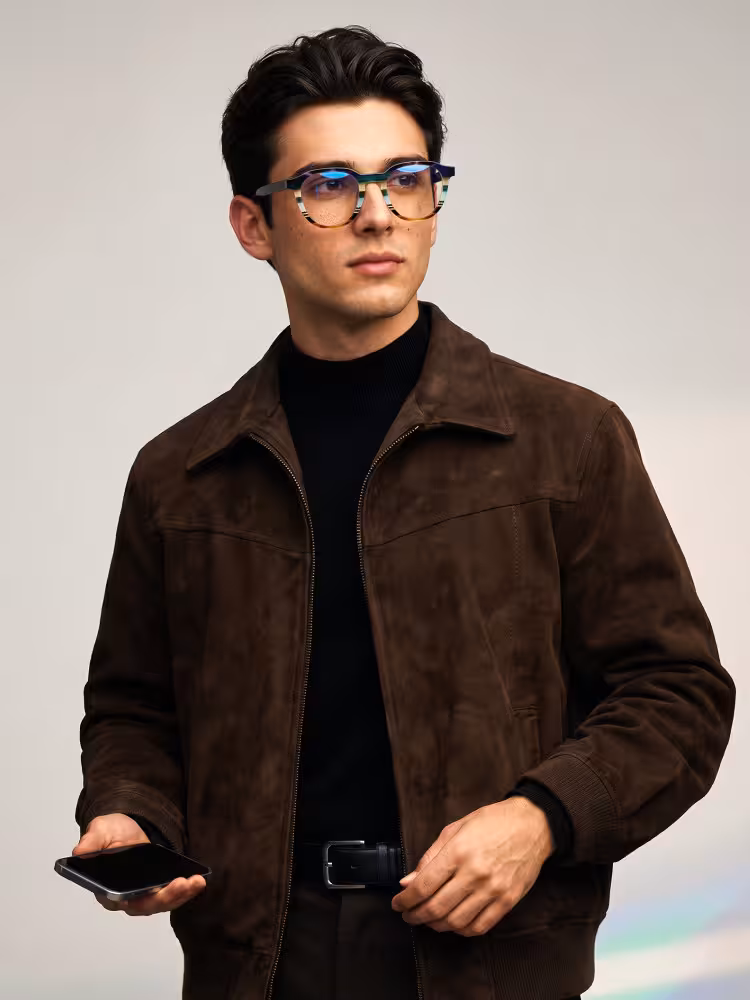 Male model wearing colorful multicolor geometric acetate eyeglasses in a brown suede jacket and black turtleneck holding a smartphone against a plain grey studio background. SPU: GA0412605