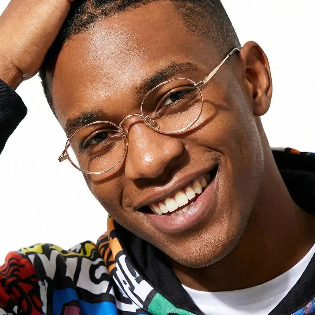 Front view of male wearing y2k gold oval eyeglasses, hand in hair, smiling, OT0282502
