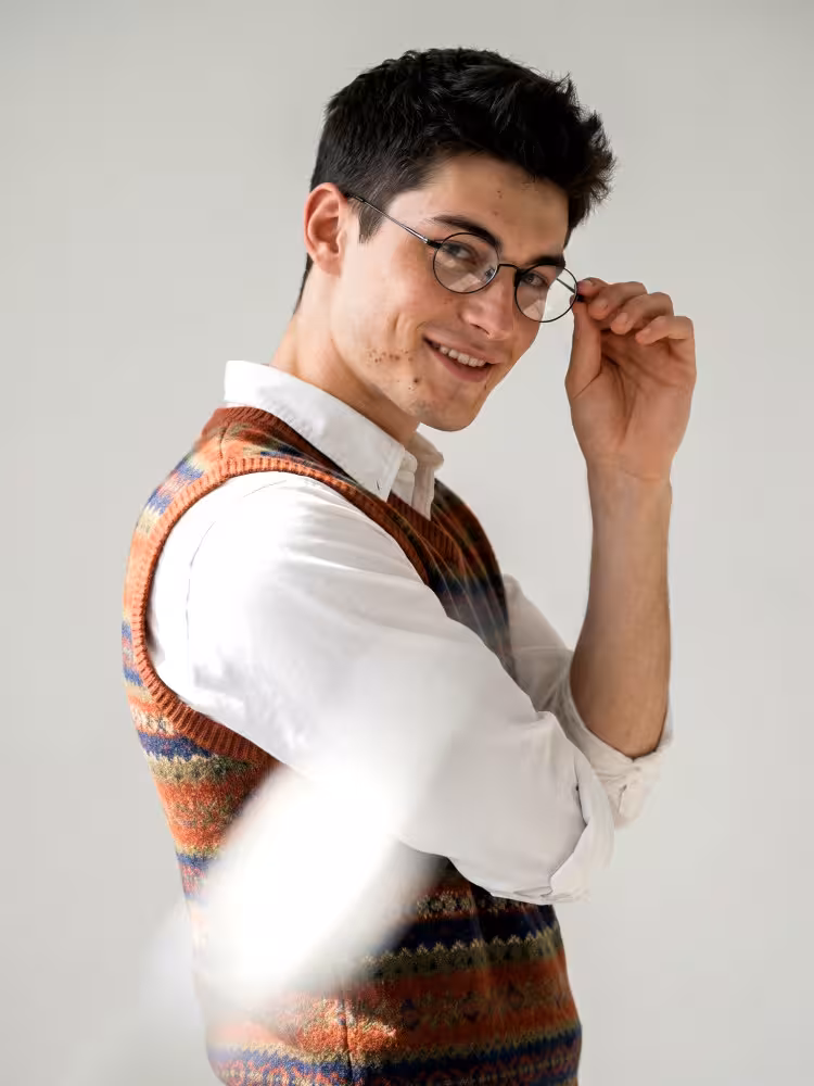 Male model wearing vintage black oval titanium eyeglasses with white shirt and retro knit vest against plain light studio background. SPU:OT0402412