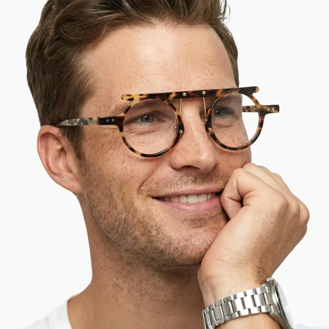 Tilted head view of male wearing tortoise round eyeglasses, resting chin on hand, natural smile, RA0012405