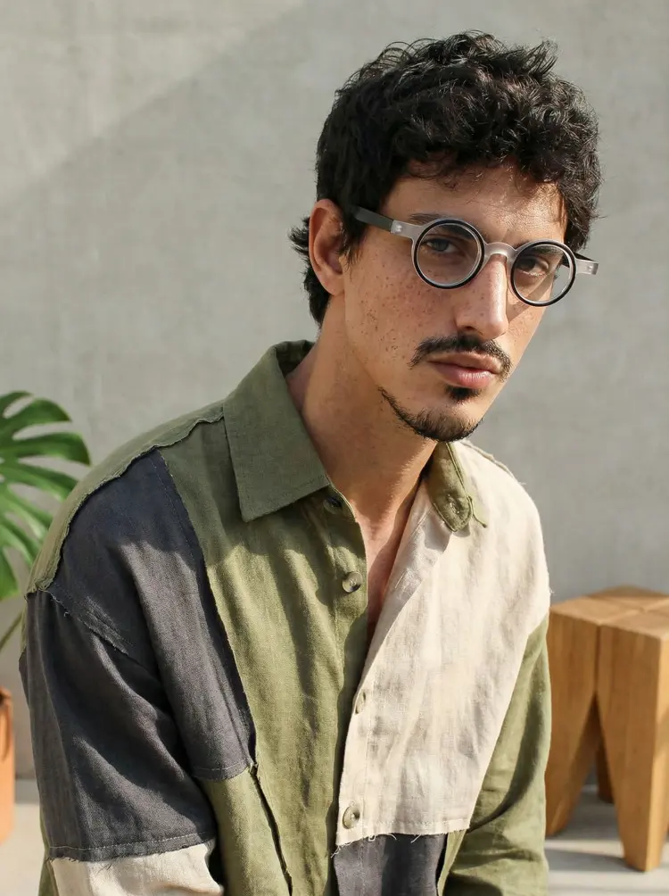 Male model wearing everyday black round acetate eyeglasses in a green and grey patchwork linen shirt against a sunny outdoor concrete wall. SPU: RA0162406