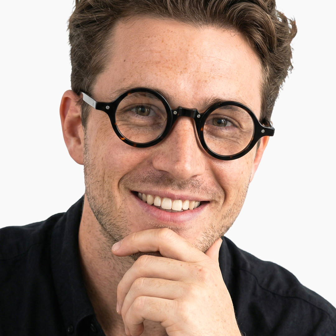 Three-quarter view of male wearing tortoise round eyeglasses, resting chin on hand, smiling, RA0182302
