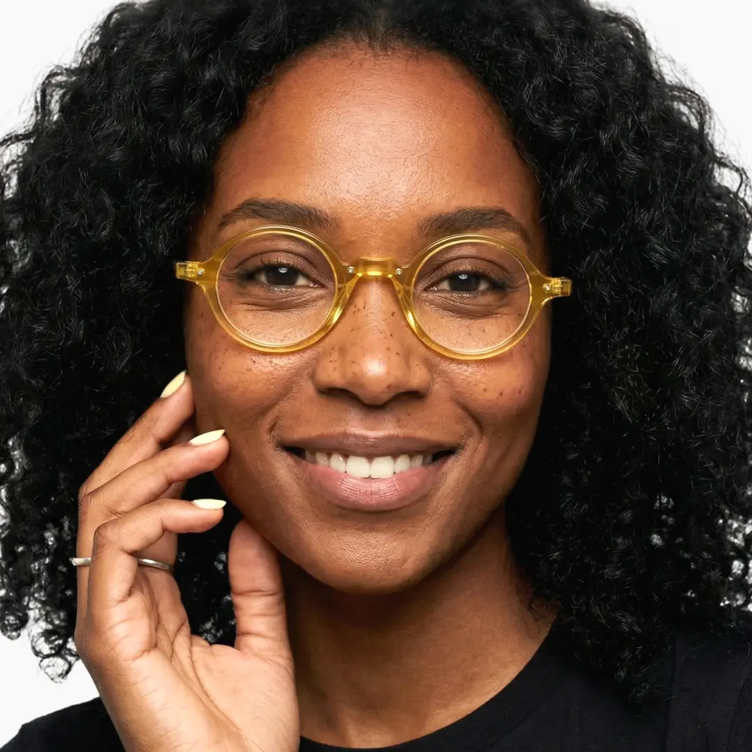Face close-up of female wearing vintage yellow round eyeglasses, hand near face, natural smile, RA0182302