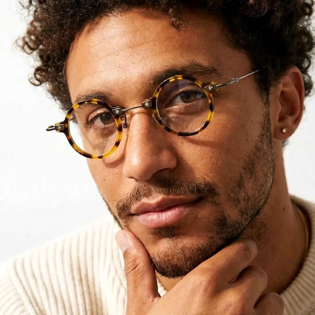 Slightly angled view of male wearing steampunk yellow tortoise round eyeglasses, hand near face, neutral expression, RA0282411