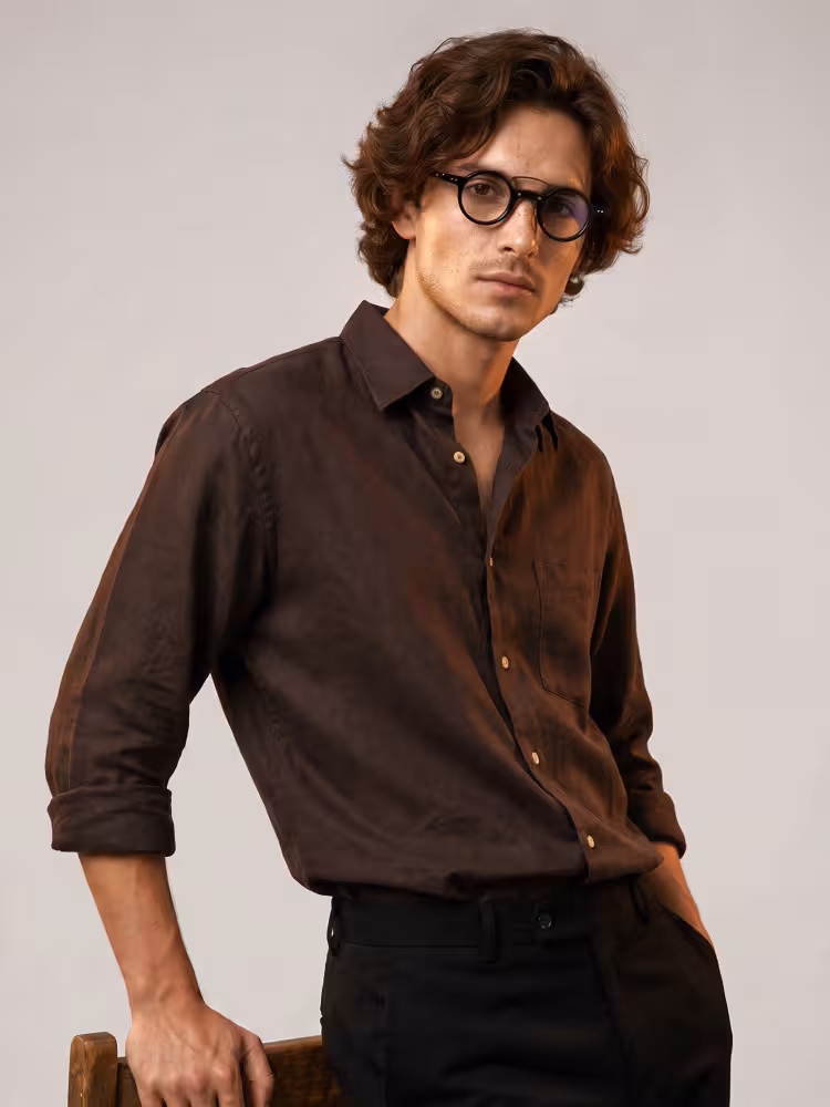 Male model wearing vintage black round acetate eyeglasses with brown linen shirt leaning on wooden chair in plain neutral studio background. SPU:RA0302310