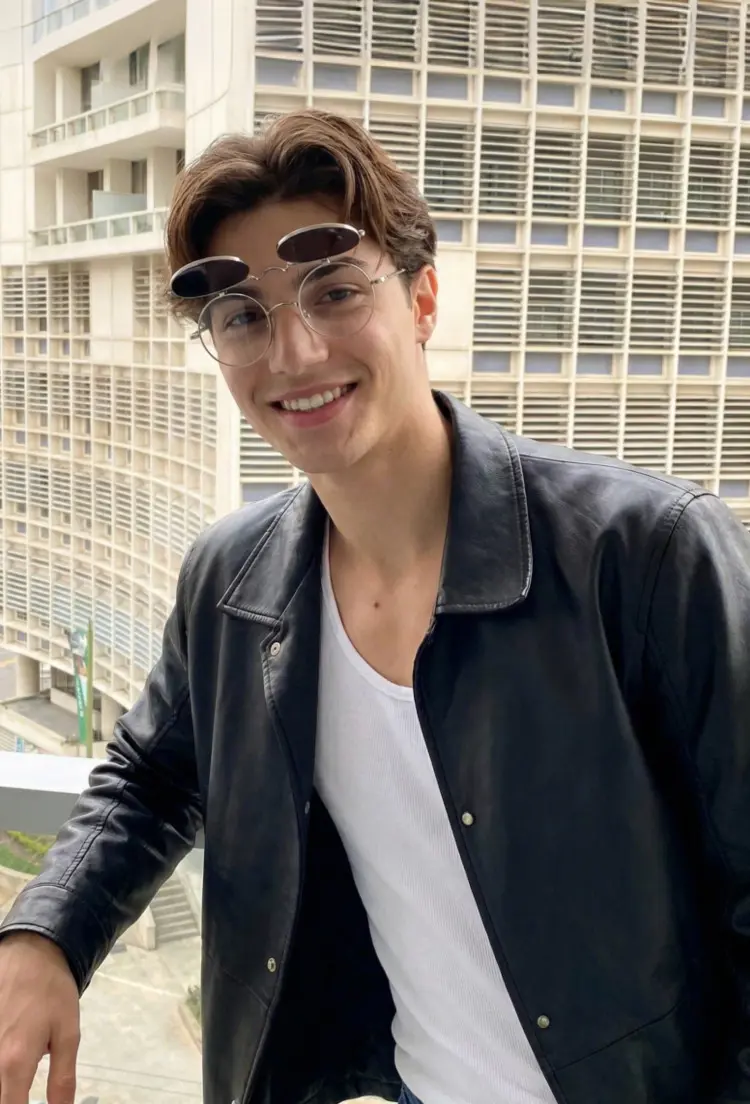 Male model wearing everyday gold round metal flip-up eyeglasses smiling in a black leather jacket with a modern city building background. SPU: RM0042601
