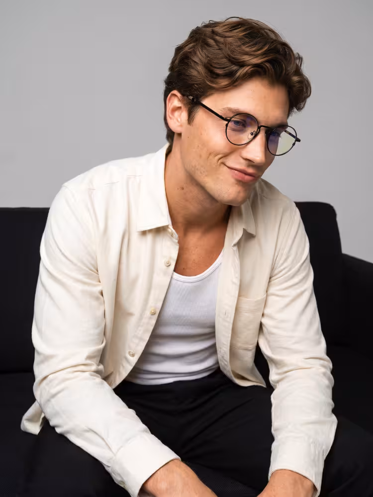 Male model wearing johnlennon black round metal eyeglasses seated on black sofa in minimalist gray studio background. SPU:RM0102407