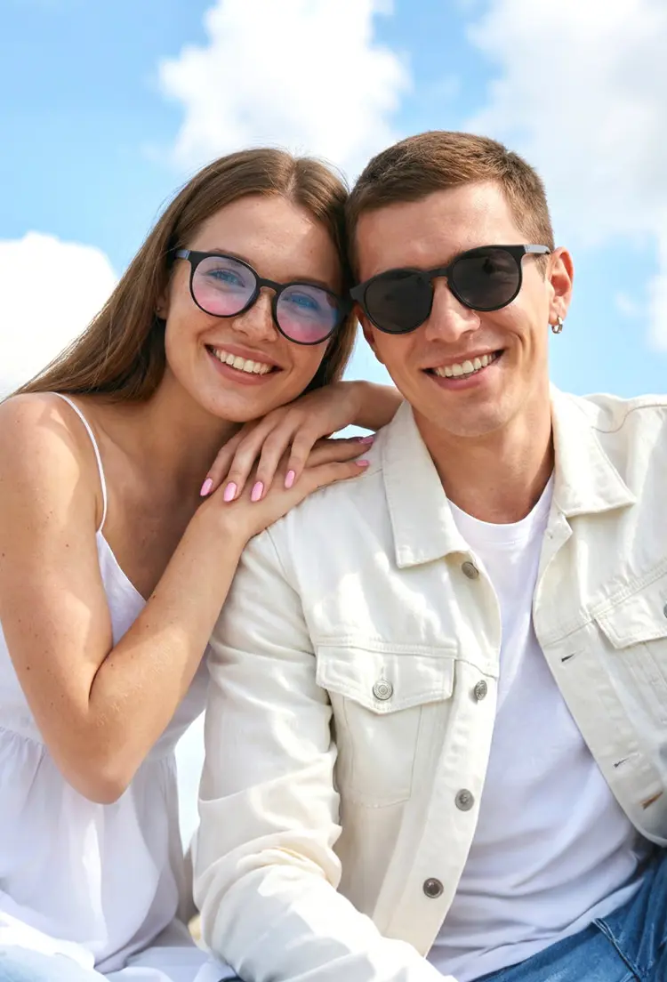 Couple model wearing magnetic black round tr90 magnetic clip-on eyeglasses set with interchangeable clip-on lenses against a bright blue sky with clouds background. SPU: RR0042512