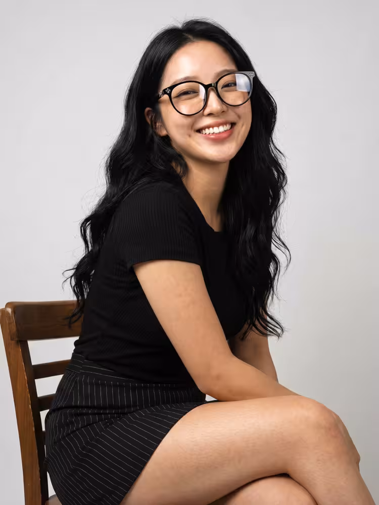 Female model wearing vaelia fashion black round tr90 eyeglasses and a black top, sitting on a wooden chair in a studio setting. SPU: RR0082409