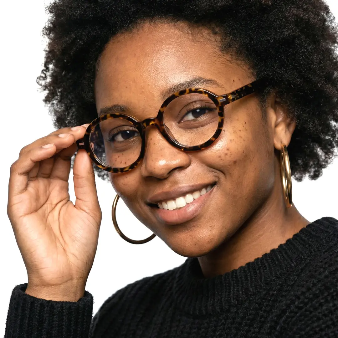 Over-the-shoulder view of female wearing brown tortoise round eyeglasses, holding glasses, smiling, RR0332302