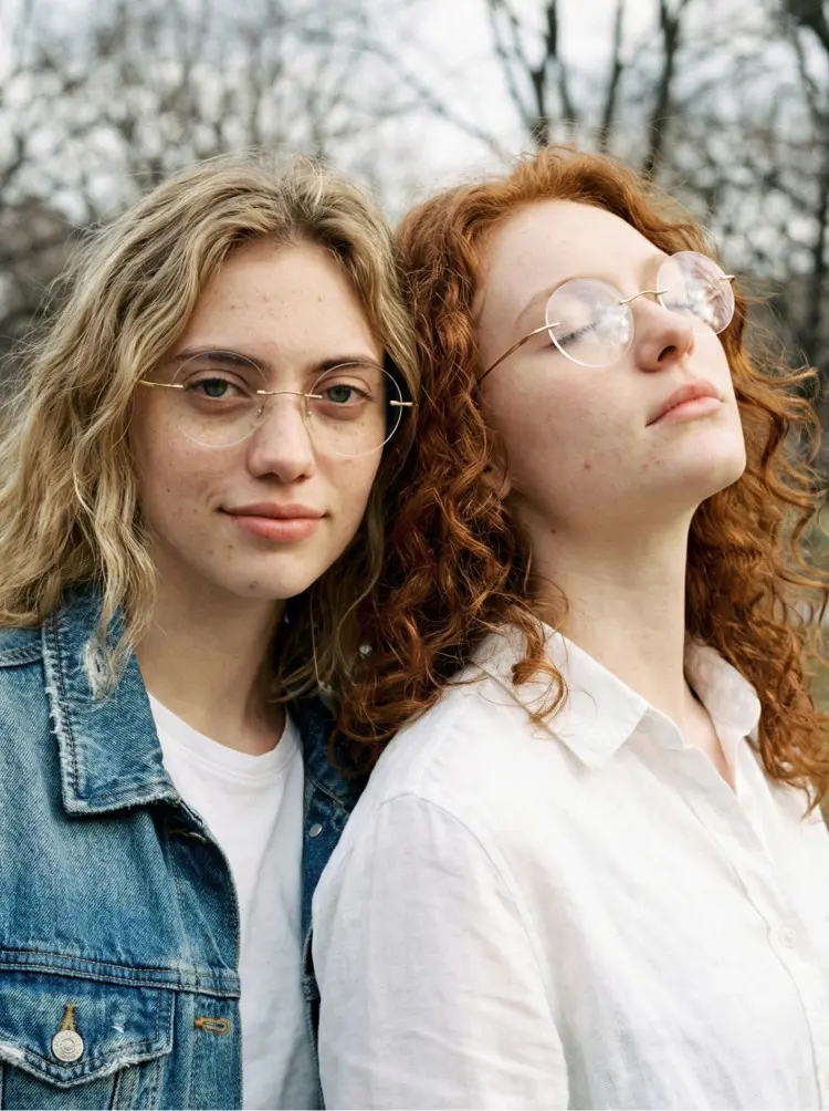 Two people model wearing vintage gold round titanium eyeglasses in an outdoor park with trees. SPU: RT0012506