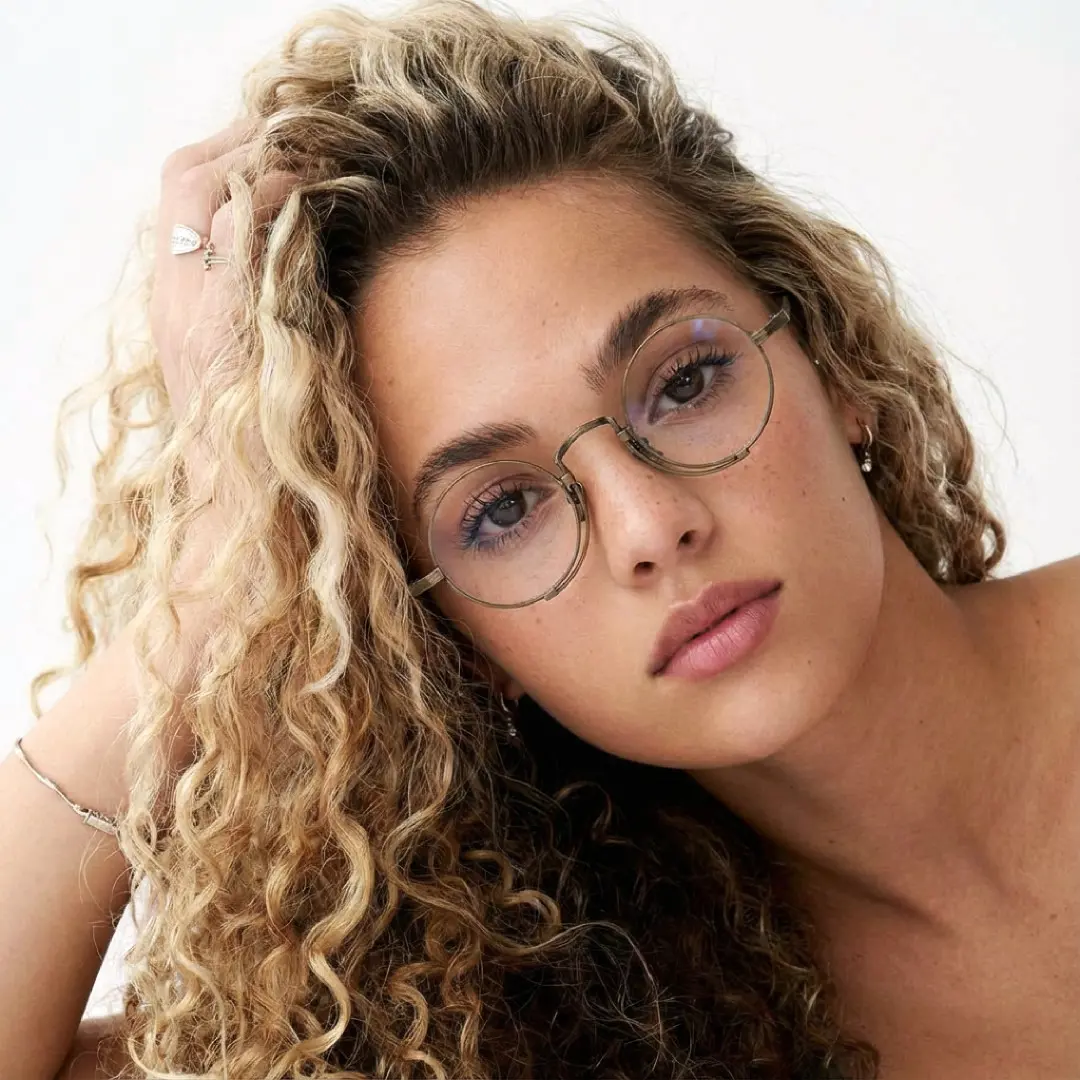 Female wearing steampunk bronze round titanium eyeglasses, neutral expression while hand in hair. SPU: RT0052304