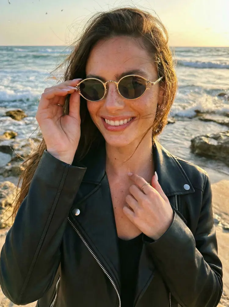 Female model wearing fashion gold oval metal sunglasses in a black leather jacket smiling on a rocky beach at sunset. SPU: OM0352601