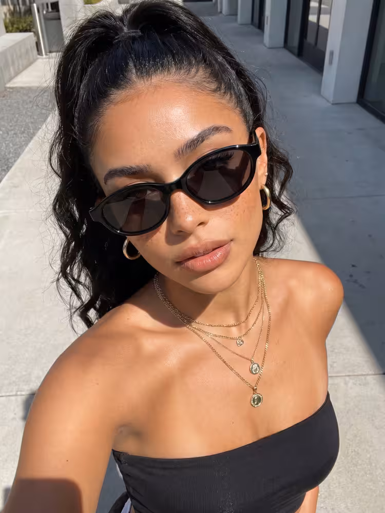 Female model wearing y2k black oval pc sunglasses taking a selfie on a sunny city sidewalk. SPU: OP0272603