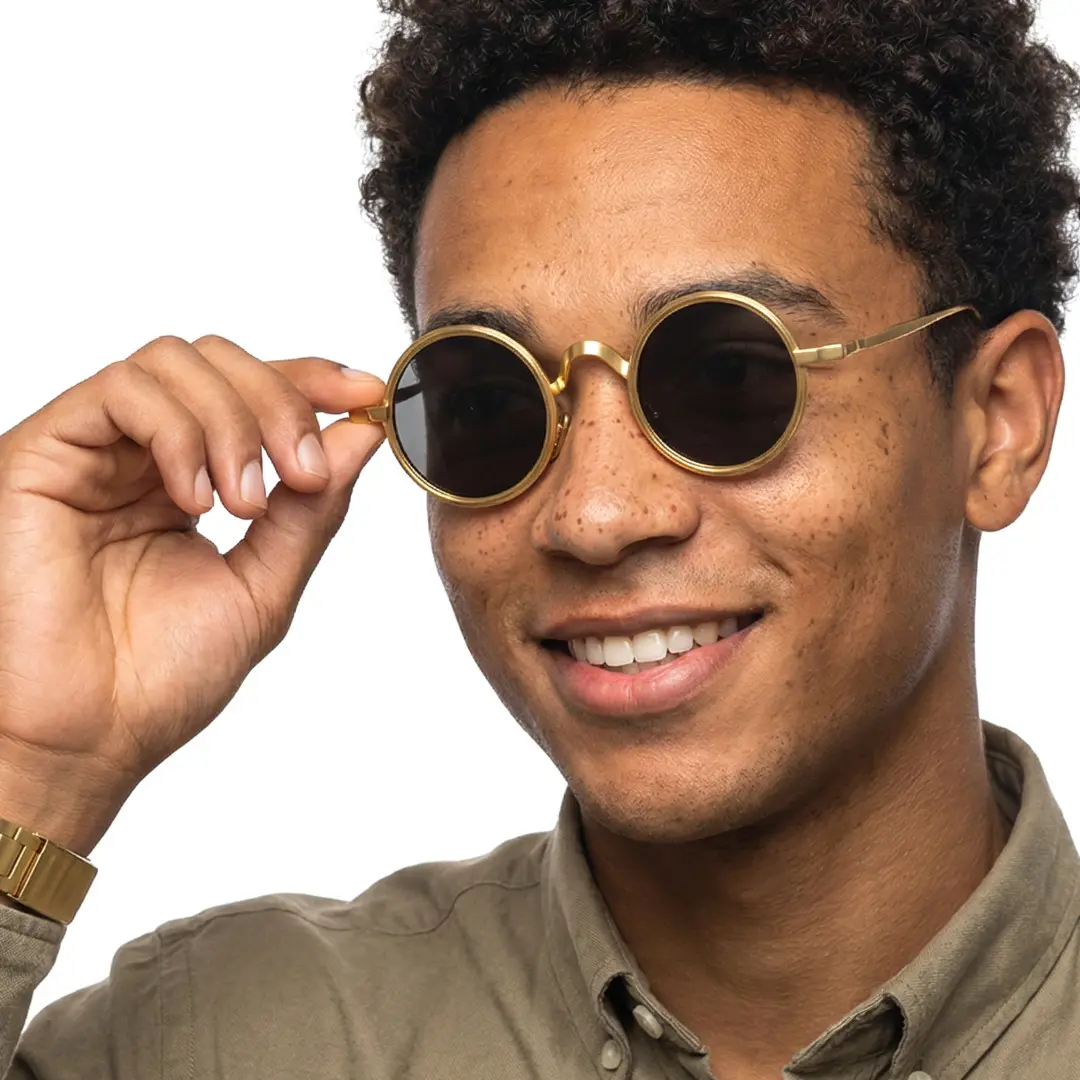 Three-quarter view of male wearing gold round sunglasses, holding glasses, soft smile, RM0162506