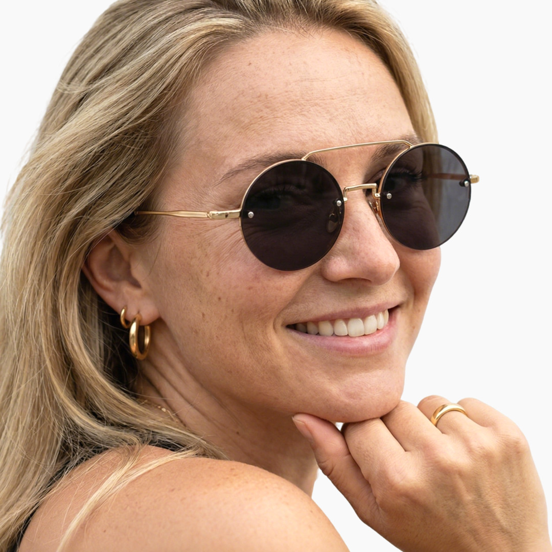 Over-the-shoulder view of female wearing vintage gold round sunglasses, resting chin on hand, natural smile, RM0252507