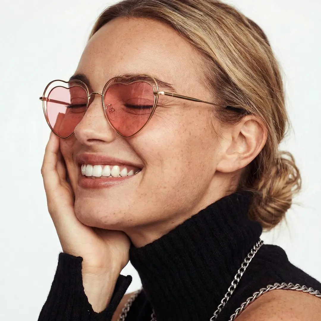 Profile view of female wearing pink heart sunglasses, resting chin on hand, smiling, UM0082506